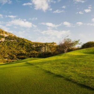 Un golfista de rosa y blanco golpea en un exuberante campo verde, con arena salpicando bajo un cielo azul con nubes: ideal para el Tour y Clínica de Golf de 1 Día entre colinas onduladas y afloramientos rocosos.