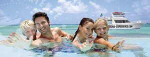 Una familia sonriente de cuatro personas, dos adultos y dos niños, nada en aguas cristalinas azules, alcanzando hacia la cámara durante su aventura 100% Island Adventure. Detrás de ellos, un barco blanco con CARIBBEAN-EXPERIENCE.COM flota en el mar turquesa.