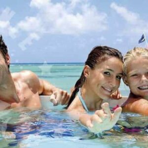 Una familia sonriente de cuatro personas, dos adultos y dos niños, nada en aguas cristalinas azules, alcanzando hacia la cámara durante su aventura 100% Island Adventure. Detrás de ellos, un barco blanco con CARIBBEAN-EXPERIENCE.COM flota en el mar turquesa.