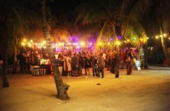 A lively nighttime beach party in Curacao, with people gathered under palm trees and colorful string lights. The sand glows warmly, creating a festive vibe as guests mingle near stalls or tables against a dark backdrop of vibrant lights.