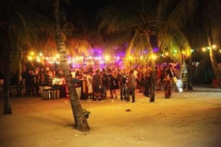 A lively nighttime beach party in Curacao, with people gathered under palm trees and colorful string lights. The sand glows warmly, creating a festive vibe as guests mingle near stalls or tables against a dark backdrop of vibrant lights.