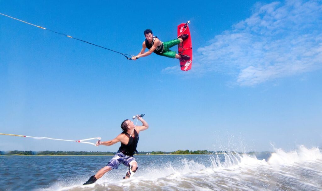 Two people are wakeboarding on a lake. One launches into an aerial trick, holding a red board and rope high above the water, while the other rides the Curacao-blue wake below, sipping from a bottle with trees and sky in the background.