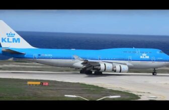 A blue and white KLM Boeing 747 aircraft is taxiing on a runway near the ocean in Curacao, with its landing gear down. The sky is clear, the sea is visible in the background, and the plane features the KLM logo on its tail and fuselage.