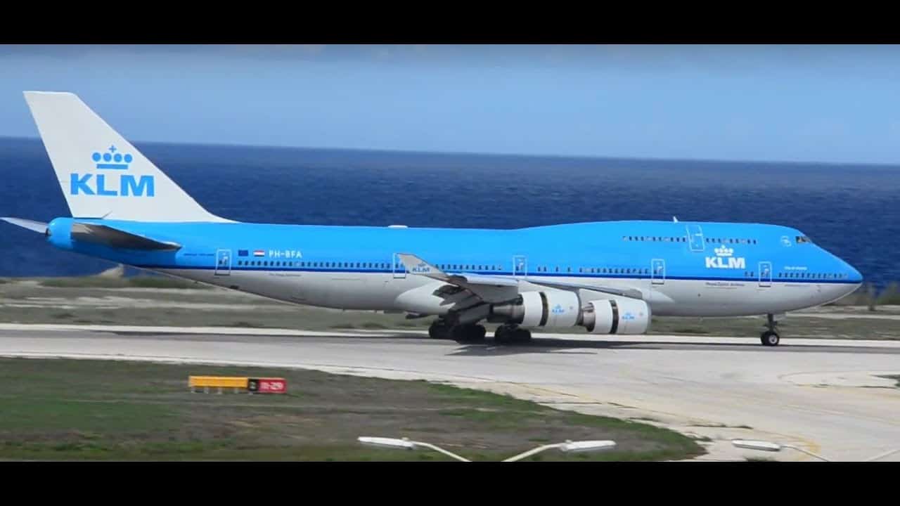 Een KLM Royal Dutch Airlines Boeing 747 met een blauw-witte kleurstelling taxiet op een landingsbaan vlakbij de oceaan in Curaçao onder een heldere hemel. Het landingsgestel van het vliegtuig is uitgeklapt en het KLM logo is zichtbaar op de staart en de romp.