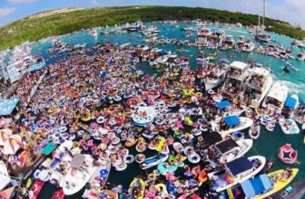 A crowded Curacao bay scene shows hundreds of people in colorful inflatable tubes and rafts gathered closely on turquoise water. Numerous boats are anchored nearby, with green hills in the background under a clear, sunny sky.