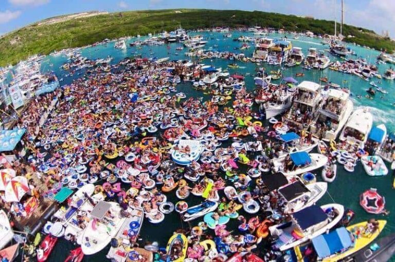 A crowded Curacao bay scene shows hundreds of people in colorful inflatable tubes and rafts gathered closely on turquoise water. Numerous boats are anchored nearby, with green hills in the background under a clear, sunny sky.