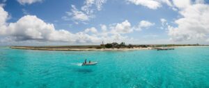 A small boat with people floats on clear turquoise water near a sandy, low-lying island in Curacao, dotted with palm trees, scattered buildings, and a dock. The bright blue sky above is filled with large, fluffy white clouds.