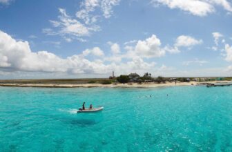 A small boat with people floats on clear turquoise water near a sandy, low-lying island in Curacao, dotted with palm trees, scattered buildings, and a dock. The bright blue sky above is filled with large, fluffy white clouds.