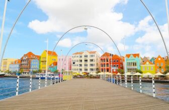 A wooden pedestrian bridge in Curacao leads toward vibrant, colorful Dutch-style buildings lining the waterfront under a bright blue sky with scattered clouds in Willemstad. Arched white lampposts curve over the bridge.
