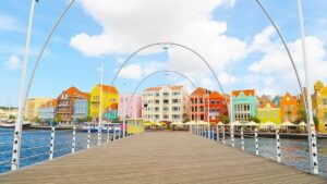 A wooden pedestrian bridge in Curacao leads toward a row of colorful Dutch colonial-style buildings along the waterfront. The sky is blue with scattered clouds, and white metal arches line the bridge. Outdoor seating and umbrellas are visible near the buildings.