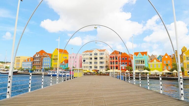 A wooden pedestrian bridge in Curacao leads toward a row of colorful Dutch colonial-style buildings along the waterfront. The sky is blue with scattered clouds, and white metal arches line the bridge. Outdoor seating and umbrellas are visible near the buildings.
