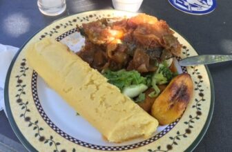 A plate with a long piece of yellow fufu, grilled fish covered in tomato-onion sauce, a slice of fried plantain, and a small salad with lettuce and cucumber brings the vibrant flavors of Curacao to your table.
