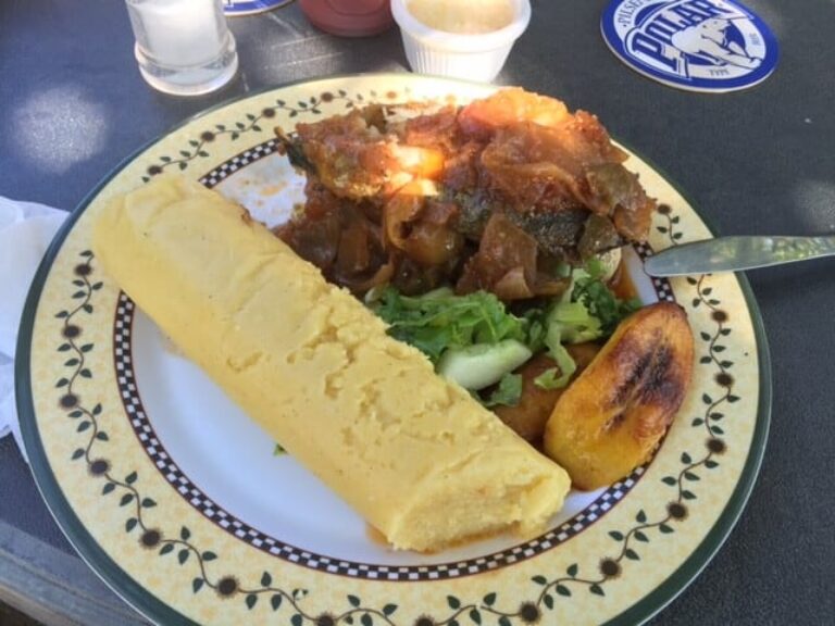 A plate with a long piece of yellow fufu, grilled fish covered in tomato-onion sauce, a slice of fried plantain, and a small salad with lettuce and cucumber brings the vibrant flavors of Curacao to your table.