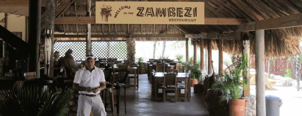 A man in a white shirt and cap smiles at the entrance of Zambezi Restaurant, an open-air spot with a thatched roof—ideal for relaxing after your African Night Special Tour at Ostrich farm.