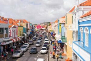 A lively Curacao street scene shows colorful colonial-style buildings with red-tiled roofs lining both sides. Crowds walk along busy sidewalks, and cars fill the road under a cloudy sky. Bright, pastel colors dominate the architecture.