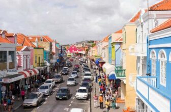 A lively Curacao street scene shows colorful colonial-style buildings with red-tiled roofs lining both sides. Crowds walk along busy sidewalks, and cars fill the road under a cloudy sky. Bright, pastel colors dominate the architecture.