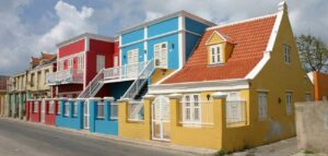 A row of colorful houses reminiscent of Curacao lines a quiet street. Painted in bright red, blue, and yellow with white shutters and orange tiled roofs, the scene is lively beneath a partly cloudy sky.