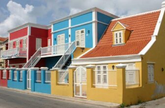 A row of colorful houses reminiscent of Curacao lines a quiet street. Painted in bright red, blue, and yellow with white shutters and orange tiled roofs, the scene is lively beneath a partly cloudy sky.