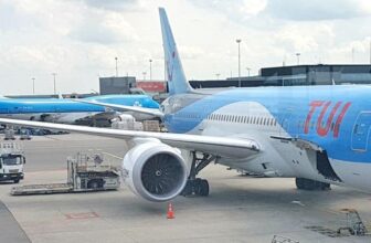 A TUI airplane is parked at an airport gate in Curacao with its engine and nose visible. Two KLM planes are seen in the background, while ground crew, luggage carts, and airport equipment are present on the tarmac under a cloudy sky.