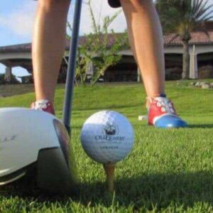 Close-up of a golf club and ball on a tee, with a golfer in red and blue shoes. Green grass, palm trees, and a large house appear under a blue sky. Featured: Advanced 1 day Package.