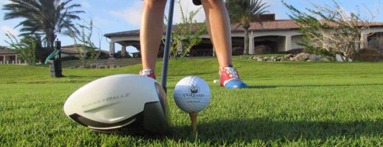 Close-up of a golf club and ball on a tee, with a golfer in red and blue shoes. Green grass, palm trees, and a large house appear under a blue sky. Featured: Advanced 1 day Package.