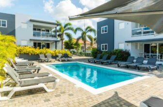 A rectangular outdoor pool surrounded by gray lounge chairs sits on a tiled patio. Modern, two-story buildings with balconies frame the scene, while palm trees and lush greenery evoke Curacao’s tropical charm under a bright, partly cloudy sky.
