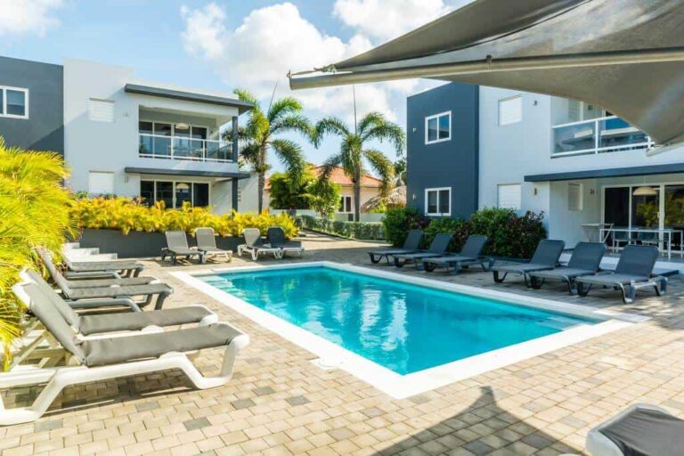 A rectangular outdoor pool surrounded by gray lounge chairs sits on a tiled patio. Modern, two-story buildings with balconies frame the scene, while palm trees and lush greenery evoke Curacao’s tropical charm under a bright, partly cloudy sky.