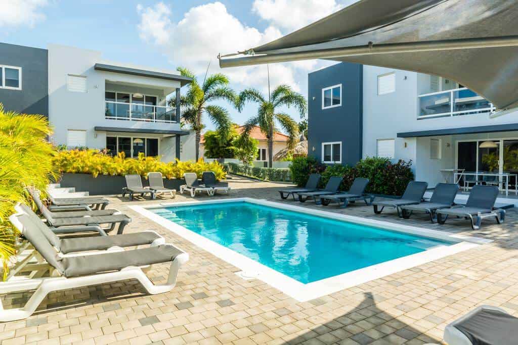 A rectangular outdoor pool surrounded by gray lounge chairs sits on a tiled patio. Modern, two-story buildings with balconies frame the scene, while palm trees and lush greenery evoke Curacao’s tropical charm under a bright, partly cloudy sky.