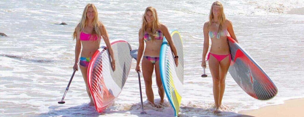 Three women enjoy Beach Hopping with SUP, standing in shallow ocean water with paddleboards and smiling as they come ashore, waves lapping their feet and sandy beach ahead.
