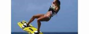 Una persona en bañador hace kitesurf sobre el océano, demostrando "Lección para principiantes 3 - Comienzo en el agua" con una tabla amarilla con arnés, elevándose bajo el cielo azul en esta dinámica escena de deportes acuáticos.