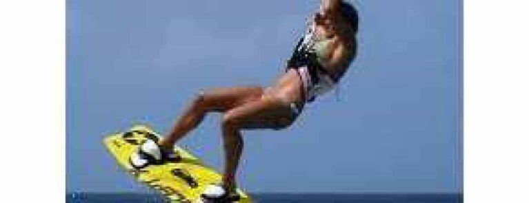 In Beginner lesson 4 – Water Start & Riding, a wakeboarder in a swimsuit and life vest soars mid-air on a yellow board against a clear blue sky, capturing the energy and excitement of the moment.