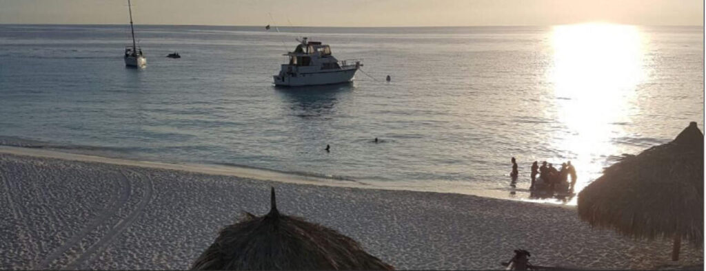 Experience Camping at Klein Curacao: Watch the sun set over calm waters with sailboats and a yacht, relax under straw-thatched umbrellas on sandy shores, and enjoy ocean reflections while standing near the water’s edge.