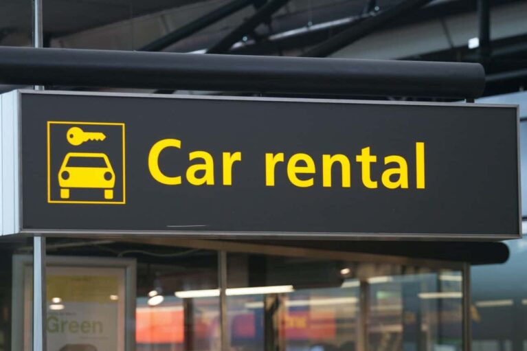 A black overhead sign with a yellow car and key icon on the left and the words “Car rental” in large yellow letters on the right, hanging inside a modern, glass-walled building in Curacao, likely an airport or transportation hub.