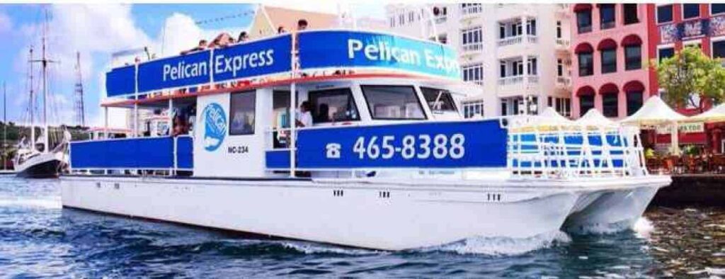 Caribbean sunset: The Pelican Express, a white and blue boat (465-8388), sails near a dock as passengers enjoy views of vibrant waterfront buildings and white tents under a partly cloudy sunset sky in the Caribbean.