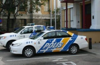 Two white Curacao police cars with blue and yellow stripes and the word POLIS parked outside a building with columns. The cars have emergency lights on top, numbers 911 and 36 visible, in an urban area with trees nearby.