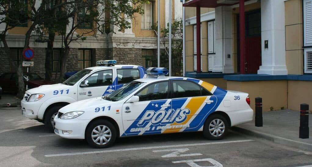 Two white Curacao police cars with blue and yellow stripes and the word POLIS parked outside a building with columns. The cars have emergency lights on top, numbers 911 and 36 visible, in an urban area with trees nearby.