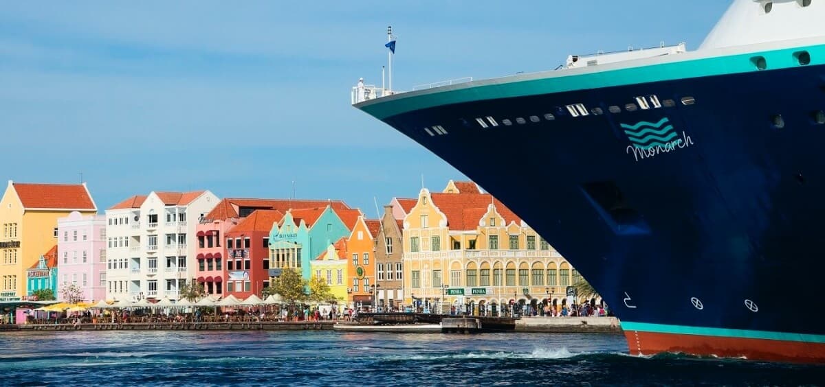 A large cruise ship’s bow dominates the right foreground near Curacao’s shoreline, lined with colorful Dutch-style buildings in pastel yellows, pinks, blues, and oranges, each topped with red-tiled roofs under a clear blue sky.