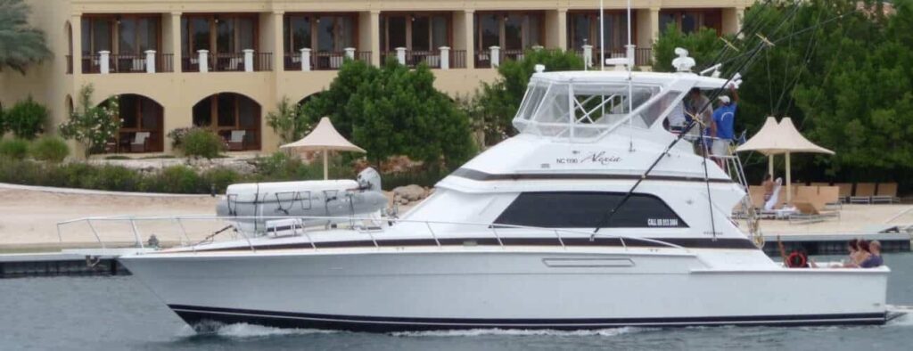A white luxury yacht from Curacao boat tours and private charters cruises near a sandy shore with guests on board, as a resort with balconies, green trees, and beige sunshades overlooks the calm water.