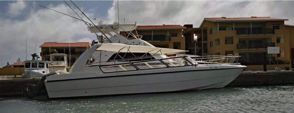 A white motor yacht with two fishing rods is docked by a concrete waterfront in Curacao. Yellow buildings with red-tiled roofs are in the background under a cloudy sky, and the Curacao watertaxi is partially visible on the left.