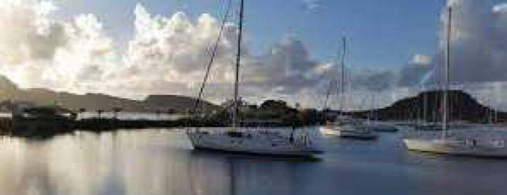 Experience a Day sail tour aboard white sailboats anchored in a marina at sunrise or sunset, with hills and a partly cloudy sky in the background. Gentle reflections of boats and clouds shimmer on the calm water’s surface.