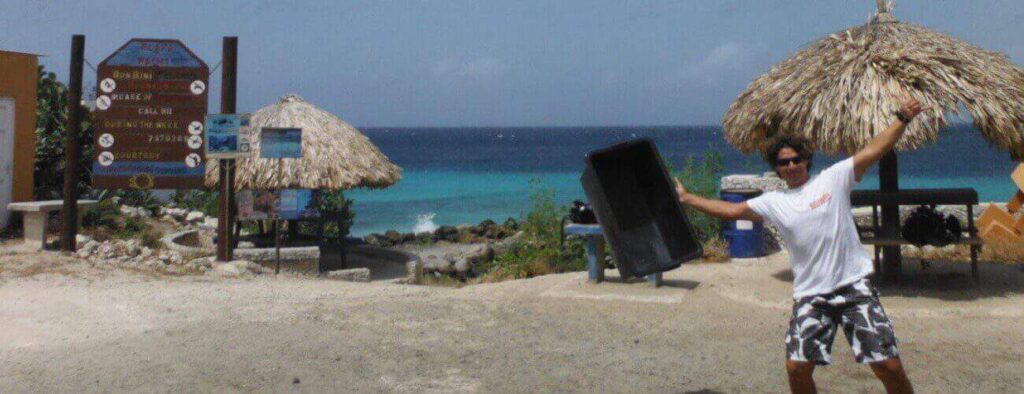 A man in sunglasses, a white tee, and patterned shorts poses with arms raised, holding "Dive in Playa Wachi" at the beach entrance, with thatched huts, turquoise ocean, and clear sky in the background.