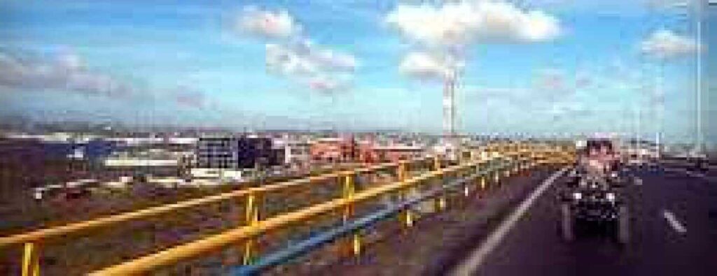 Op een heldere dag rijden twee mensen op een motor over een verhoogde East Side-snelweg met gele en blauwe vangrails. Industriële gebouwen en een gedeeltelijk bewolkte lucht zijn op de achtergrond te zien tijdens de East Side rondleiding.