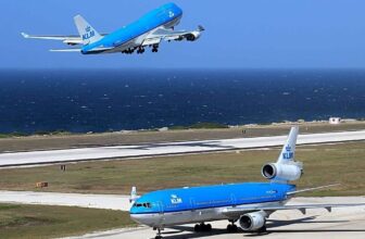 A KLM blue and white Boeing 747 is taking off over a runway near the ocean in Curacao, while a KLM McDonnell Douglas MD-11 taxis on the ground below. Both planes display the KLM logo against a bright blue sky and deep blue sea.