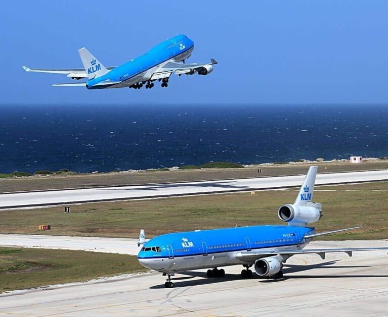 A KLM blue and white Boeing 747 is taking off over a runway near the ocean in Curacao, while a KLM McDonnell Douglas MD-11 taxis on the ground below. Both planes display the KLM logo against a bright blue sky and deep blue sea.
