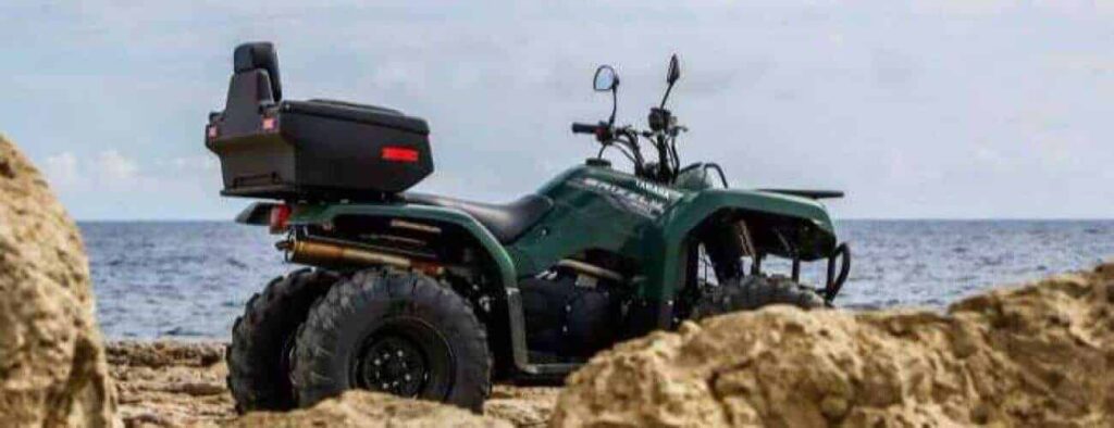 De "Halve dag ATV-avontuur oostkant" biedt ritten op een groene ATV met zwarte opbergbox langs de rotsige kust en oceaan, onder een bewolkte hemel-perfect decor voor jouw halve dag vol avontuur.
