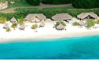 Aerial view of a Curacao tropical beach with turquoise water, white sand, and thatched-roof huts along the shore, surrounded by lush green trees. People relax under umbrellas while a small boat floats near the water’s edge.