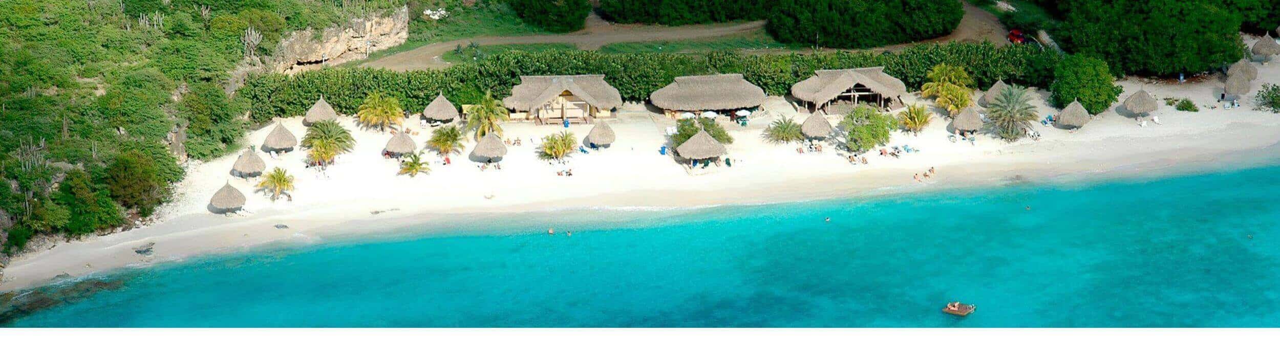 Aerial view of a Curacao tropical beach with turquoise water, white sand, and thatched-roof huts along the shore, surrounded by lush green trees. People relax under umbrellas while a small boat floats near the water’s edge.
