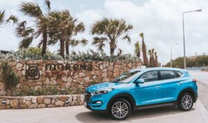 A blue SUV is parked on a road next to a stone wall with the words Morena Resort and a circular logo in Curacao. Palm trees sway behind the wall under a partly cloudy sky, while streetlights line the road on the right.