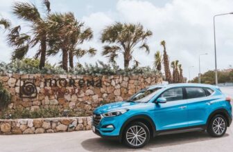 A blue SUV is parked on a road next to a stone wall with the words Morena Resort and a circular logo in Curacao. Palm trees sway behind the wall under a partly cloudy sky, while streetlights line the road on the right.