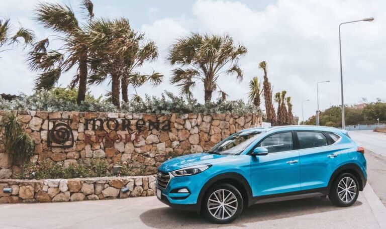 A blue SUV is parked on a road next to a stone wall with the words Morena Resort and a circular logo in Curacao. Palm trees sway behind the wall under a partly cloudy sky, while streetlights line the road on the right.